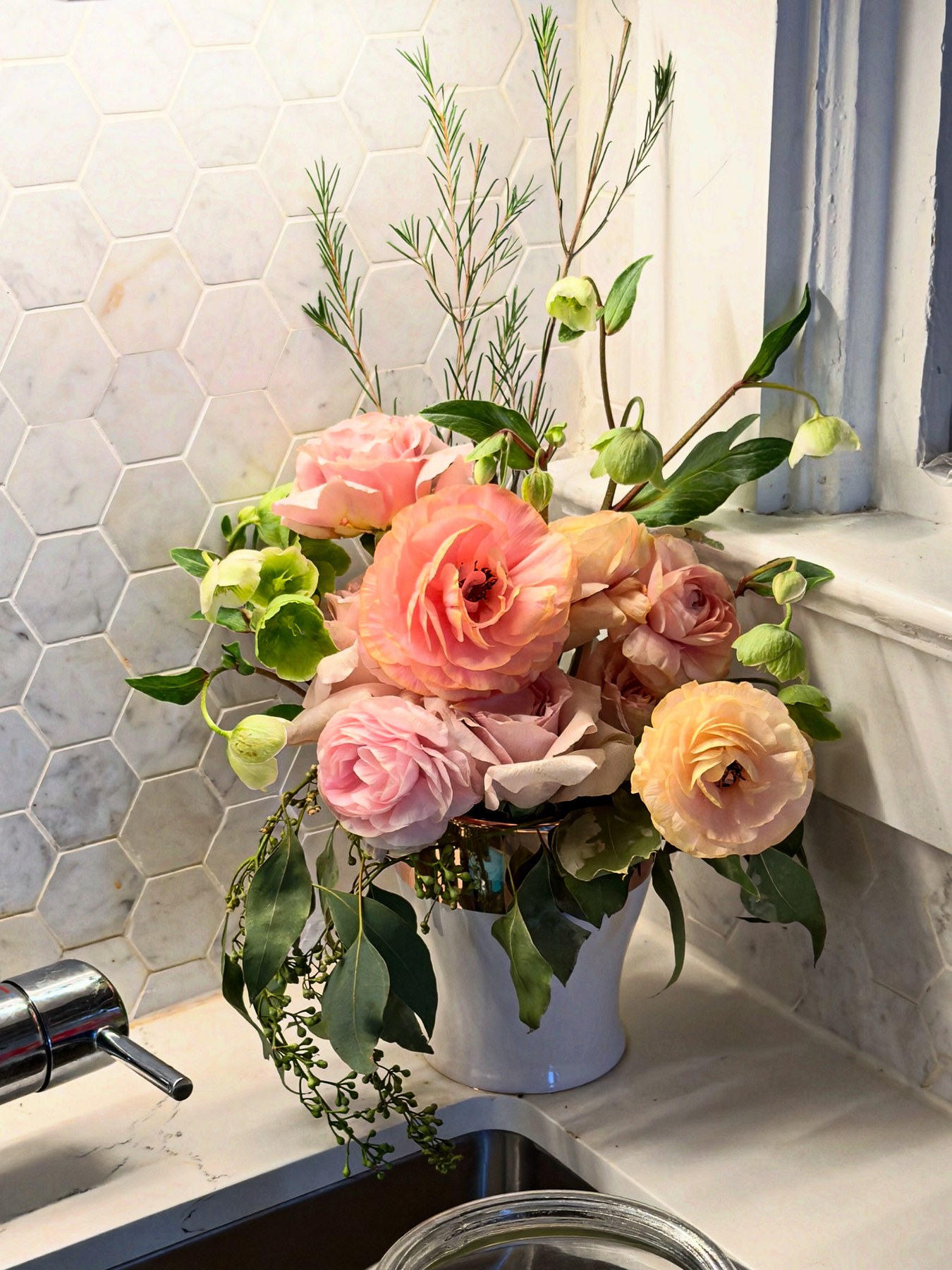 Blush rose and ranunculus arrangement with rosemary in white vessel by kitchen sink
