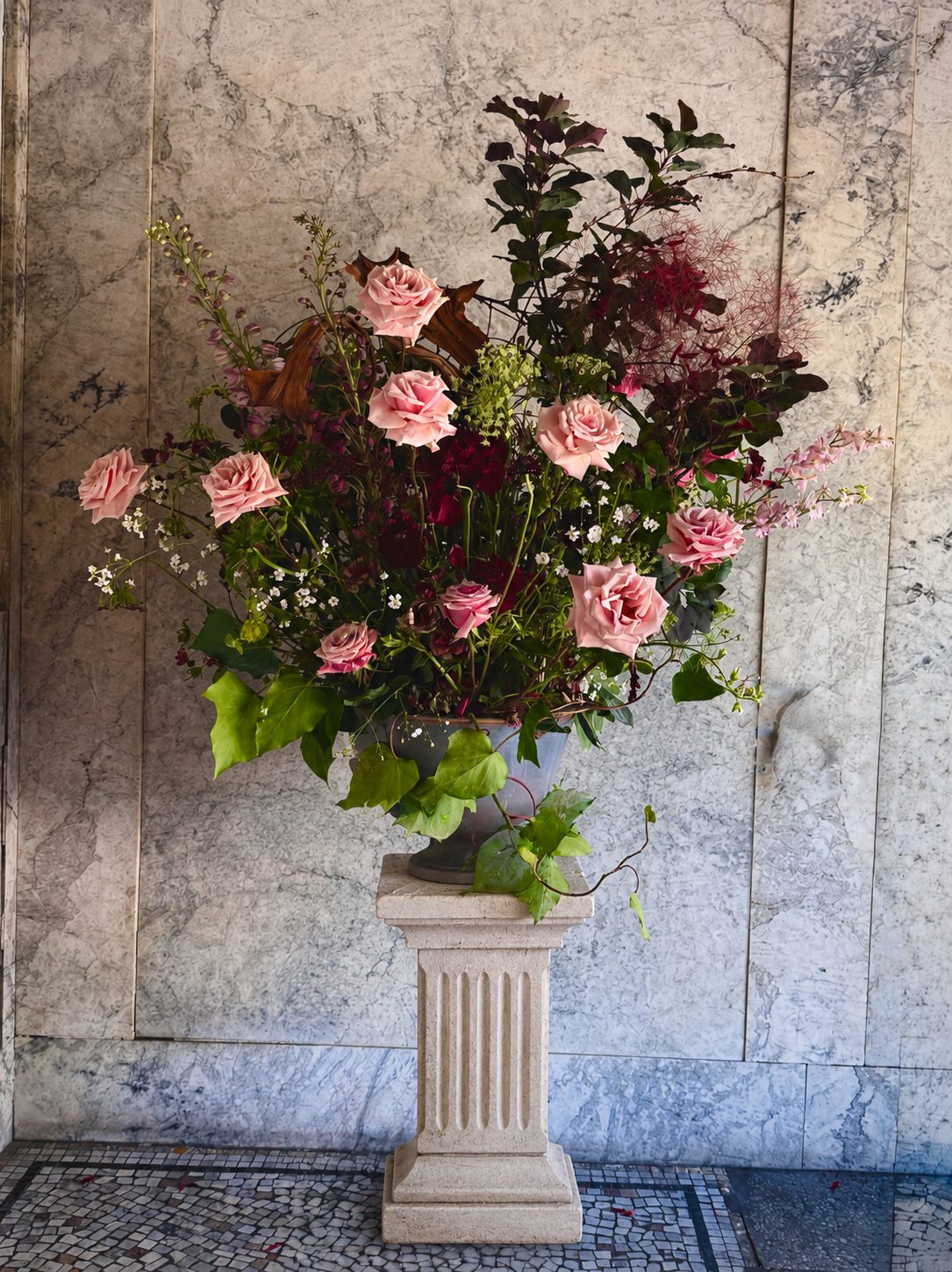 Blush roses with smoke bush in tall urn against marble wall