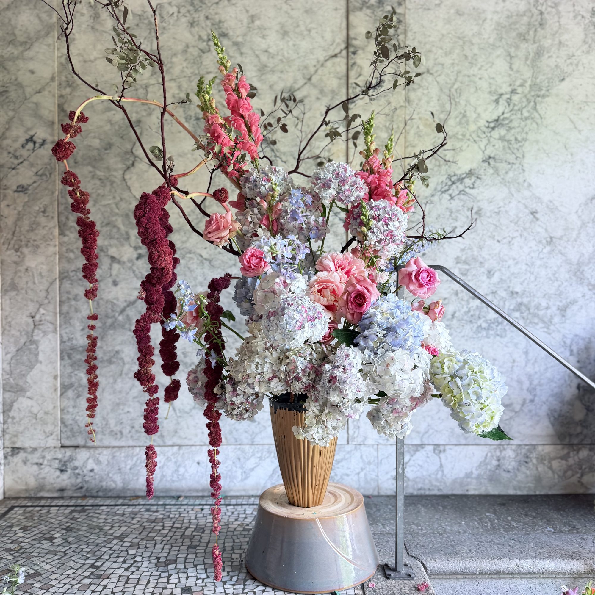 Lavender hydrangeas with trailing amaranthus in a tall urn against marble wall