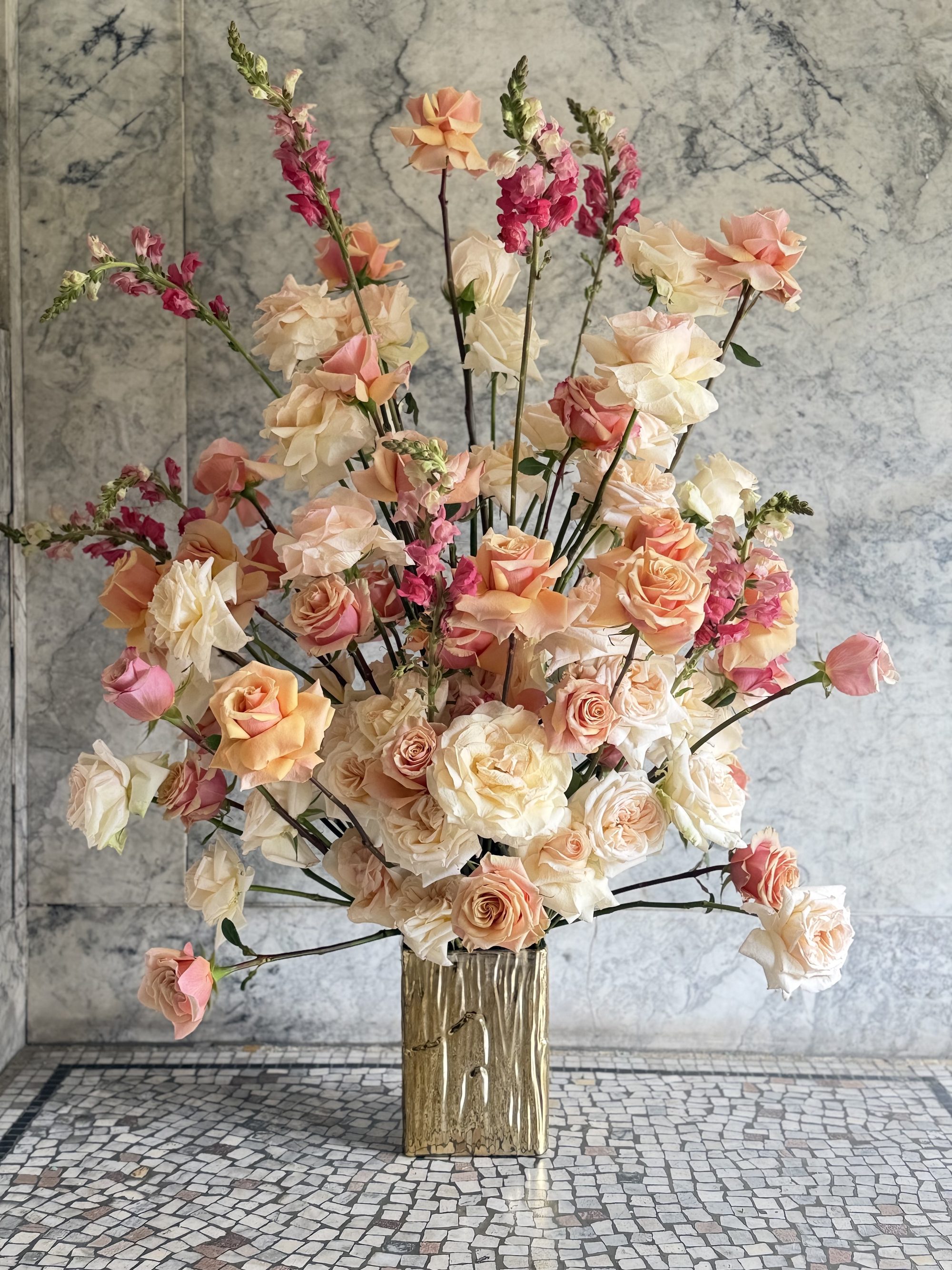 Peach roses and snapdragons in gold vessel against marble wall
