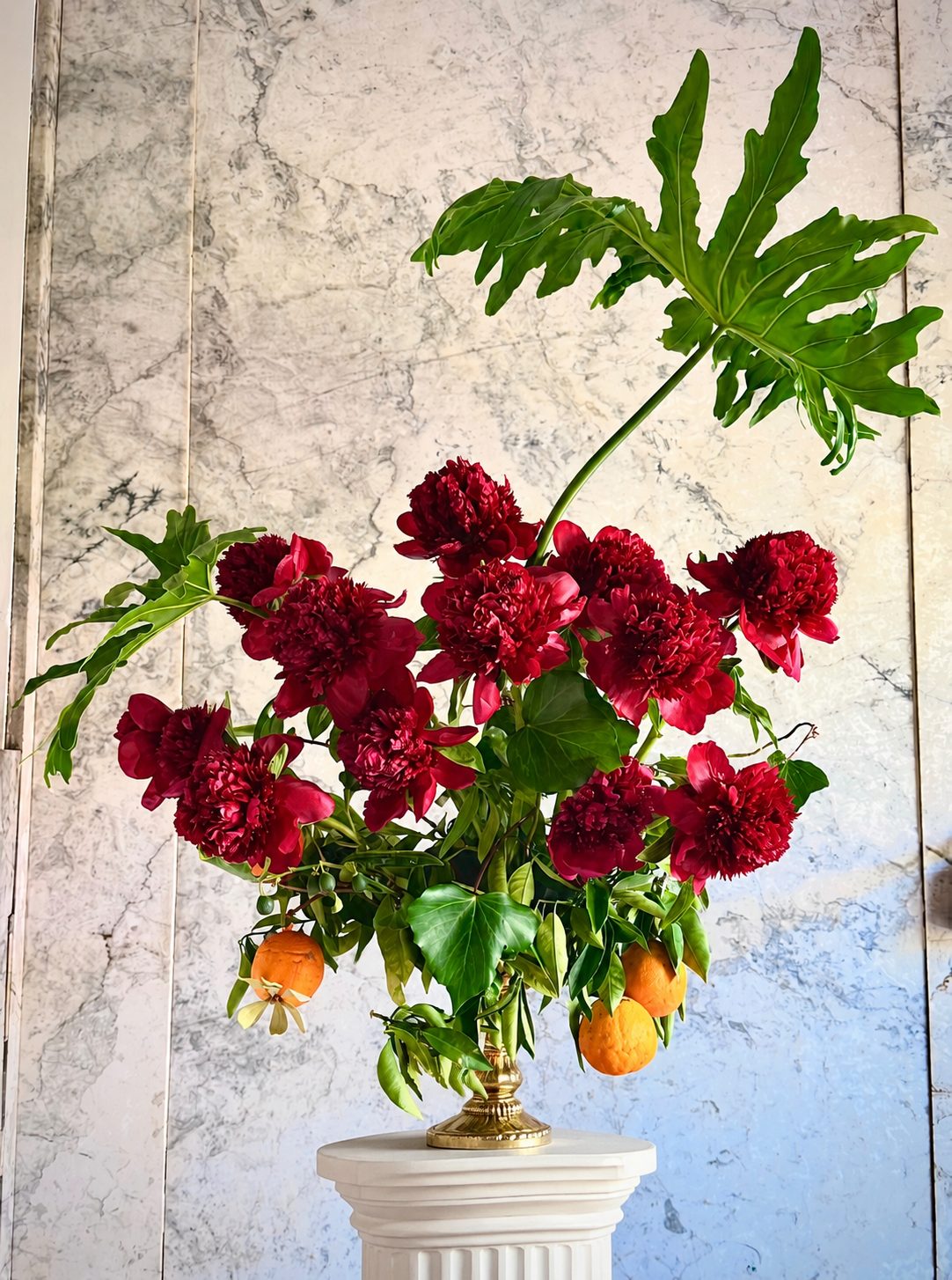 Red peonies with monstera leaves on pedestal against marble wall