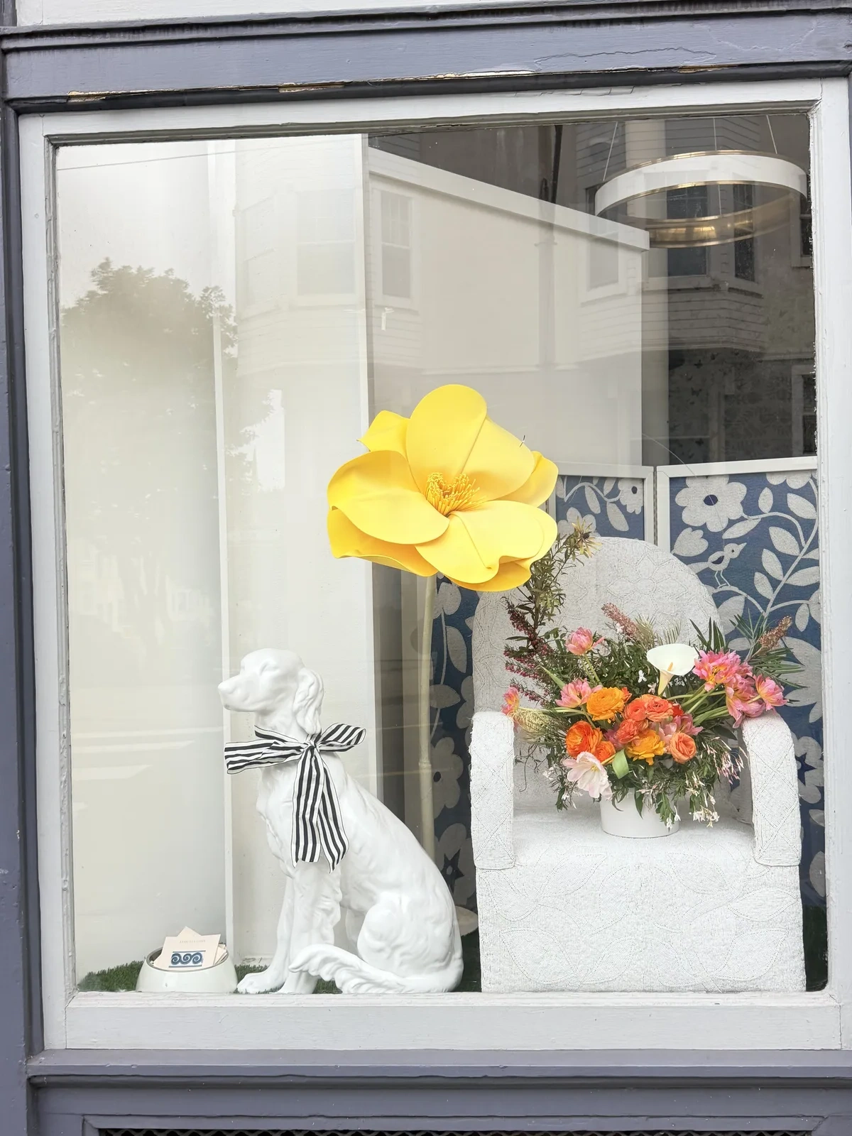 Window installation by Lovely Coven for Amy Weaver — oversized yellow paper bloom, white dog statuette with striped ribbon, and a low arrangement of orange ranunculus and pink garden roses.