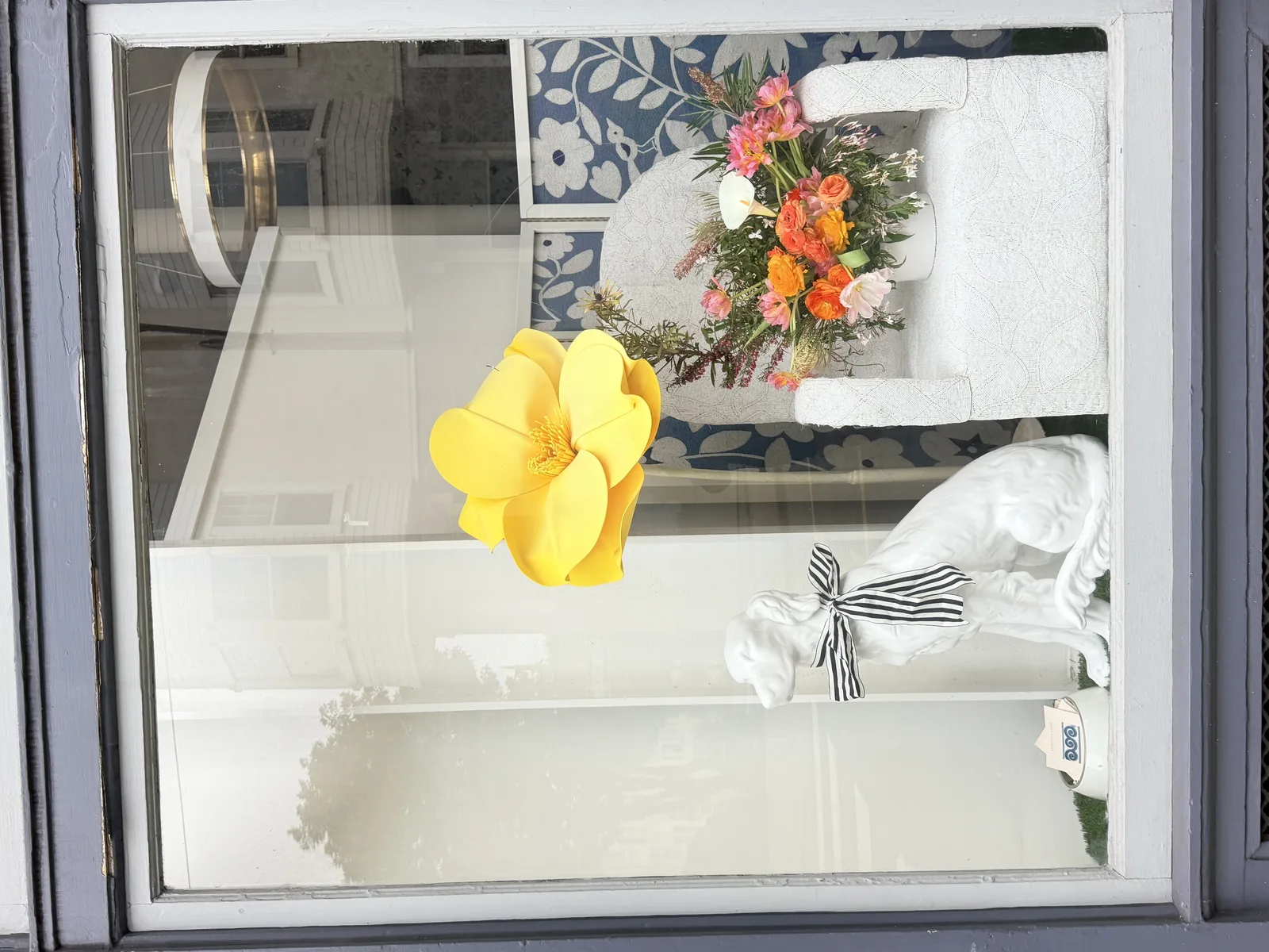 Window installation by Lovely Coven for Amy Weaver — oversized yellow paper bloom, white dog statuette with striped ribbon, and a low arrangement of orange ranunculus and pink garden roses.