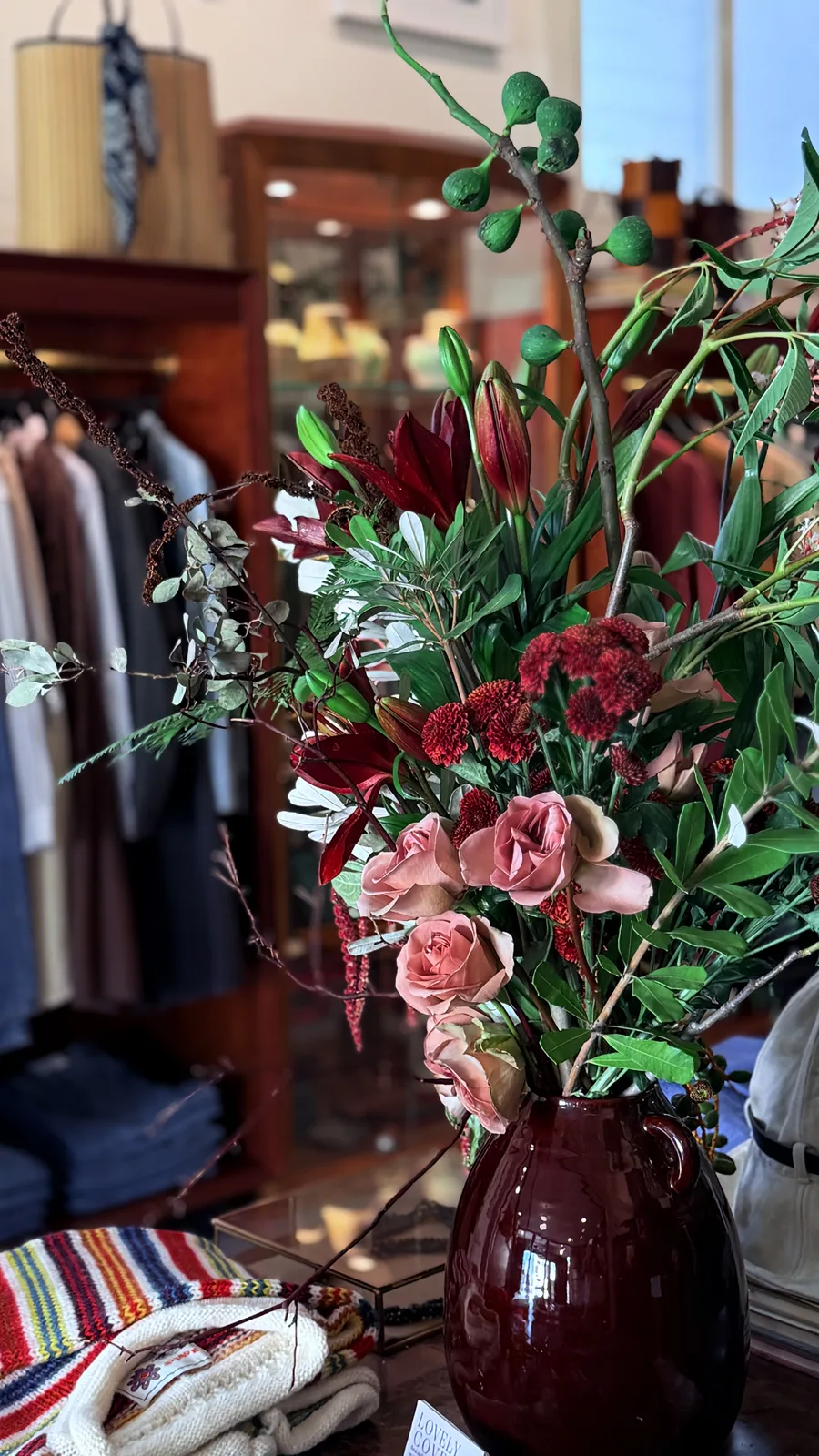 Lovely Coven arrangement at Presidio Post — burgundy pitcher with deep red lilies, garden roses in dusty rose, eucalyptus pods, and trailing greenery, set on a wooden surface inside the shop.