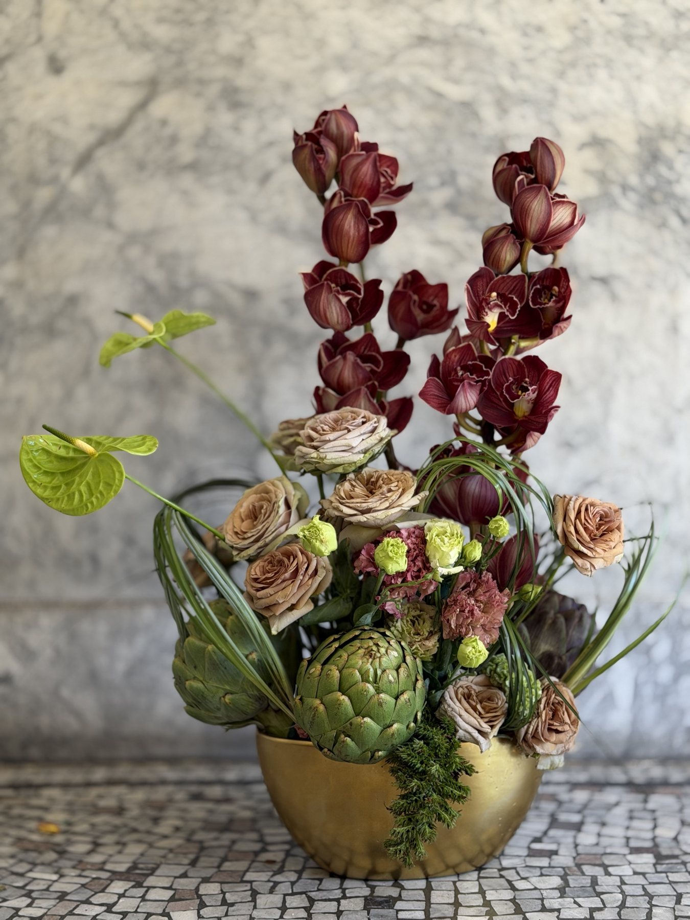 Two cymbidium spikes rise above artichokes and toffee roses in a gold bowl, with a lime anthurium leaning outward