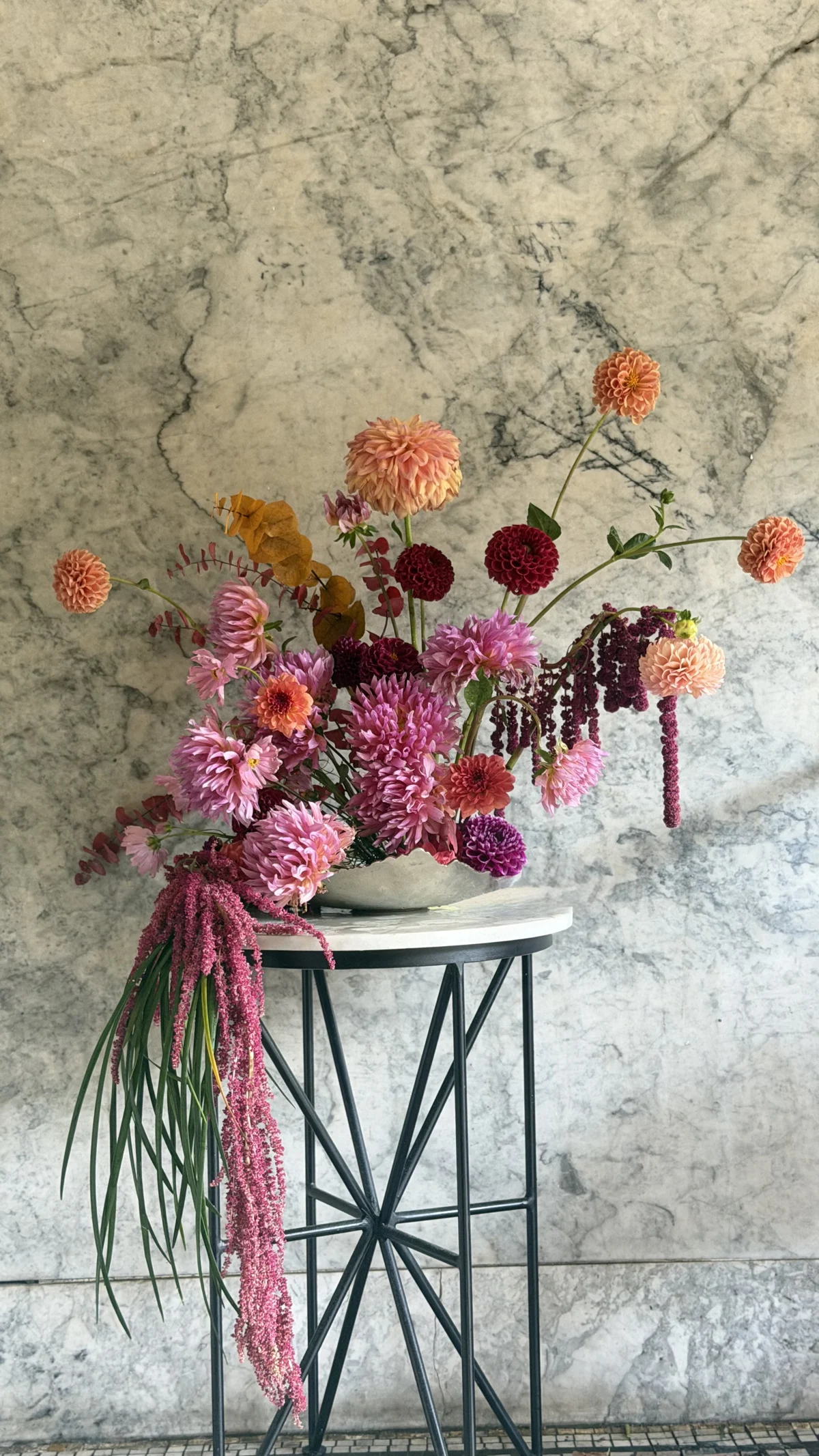 Coral and burgundy dahlias rise above lavender-pink chrysanthemums, copper-toned foliage and trailing amaranthus in a pale ceramic vessel on a tall black stand