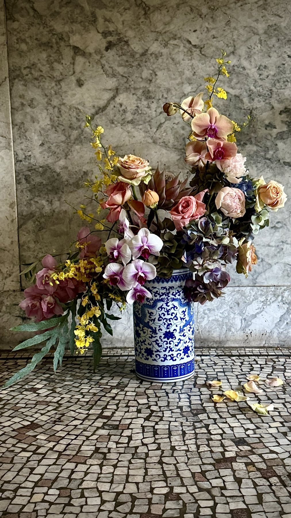 Yellow oncidium sprays above phalaenopsis, roses, peonies and hydrangea in a dragon vessel, with fallen petals below