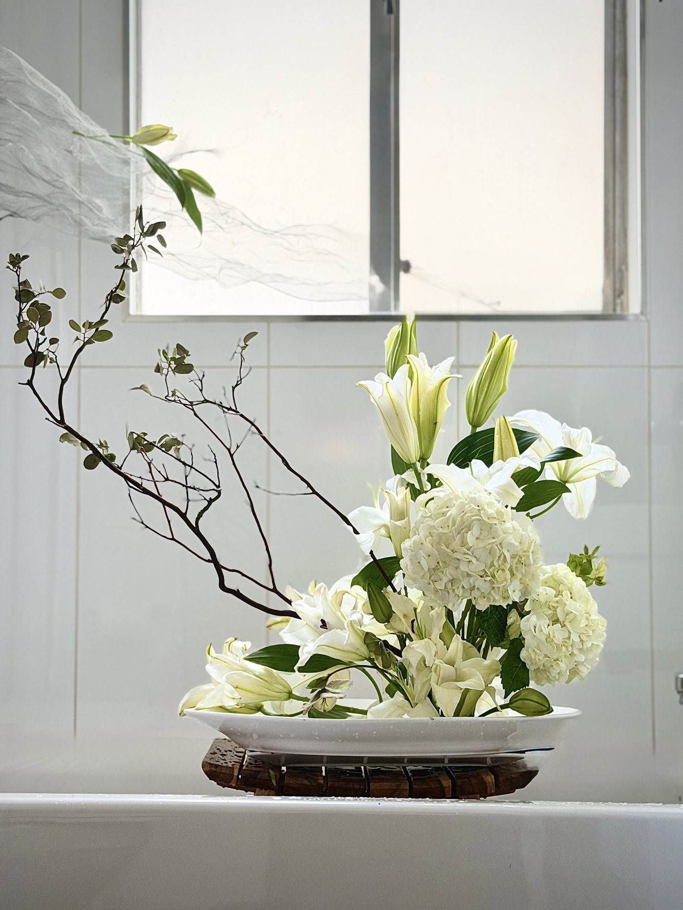 White Oriental lilies and hydrangea with a single bare branch in a low white dish