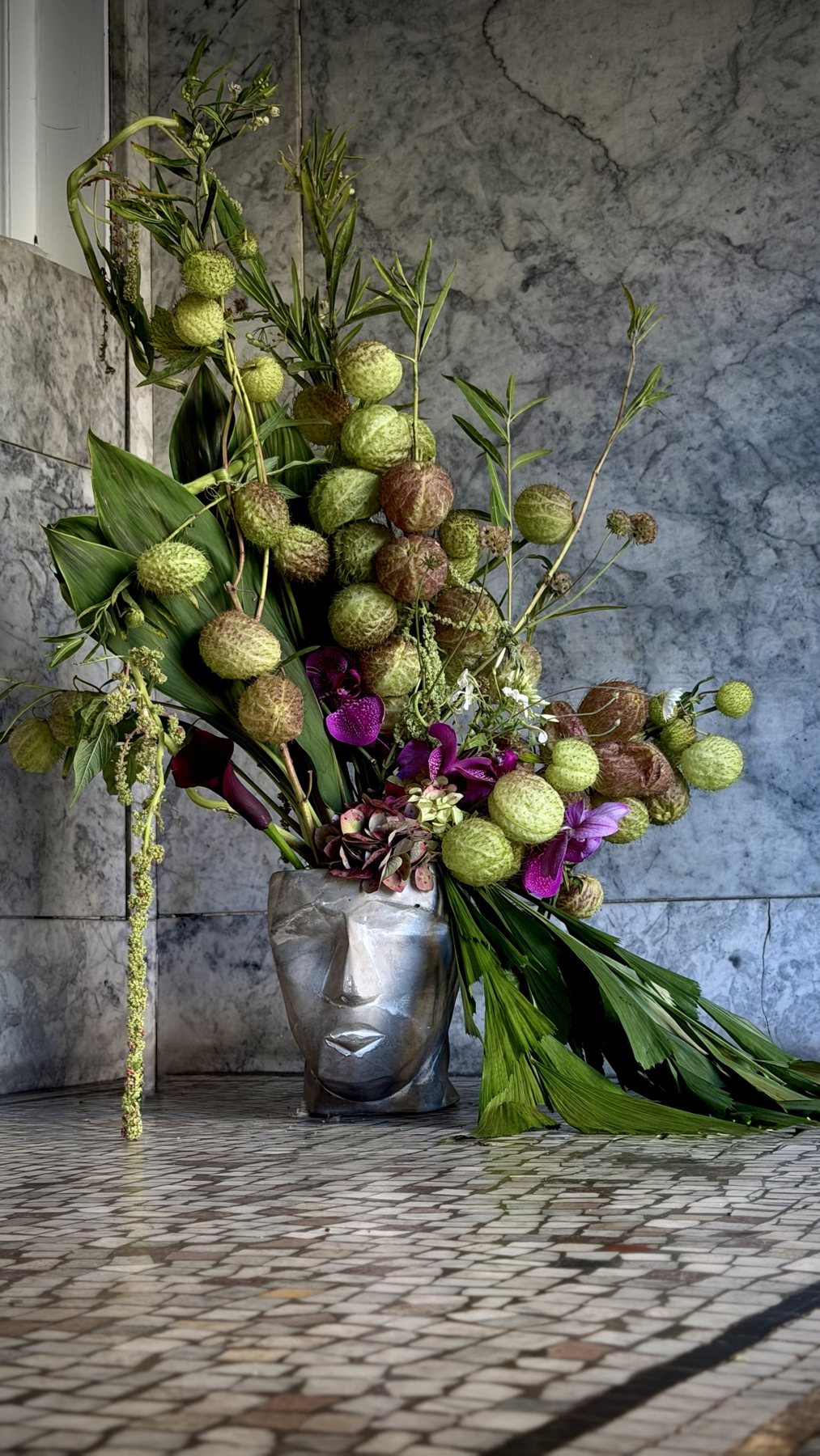Balloon milkweed pods and orchids rise from a silver sculpted head, with palm leaves trailing across the floor