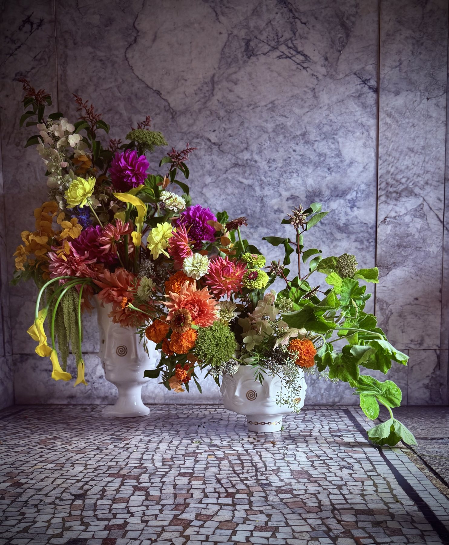 Two face vessels hold dahlias, marigolds, zinnias and callas, with geranium trailing from the lower arrangement