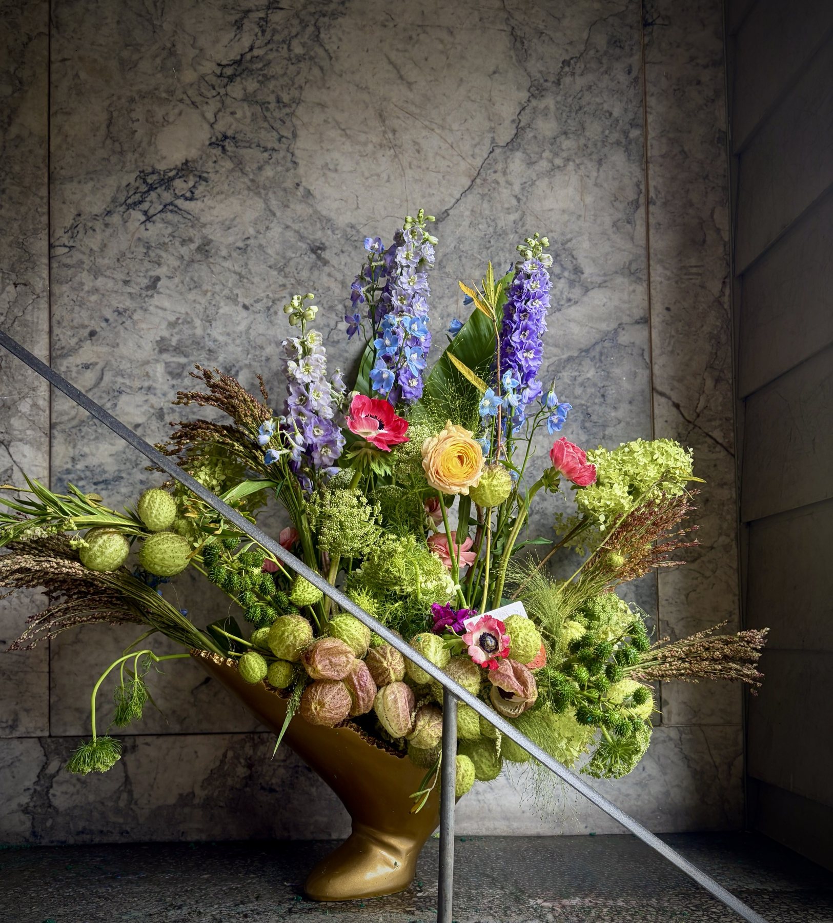 Delphinium, anemones, ranunculus, milkweed pods and hydrangea in a gold vessel beside a stair rail