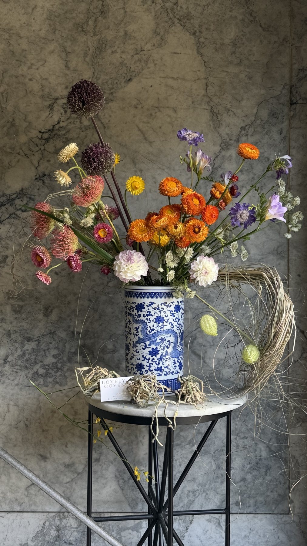 Strawflowers, allium and dahlias arranged beneath a loose loop of dried grass in a blue-and-white vessel