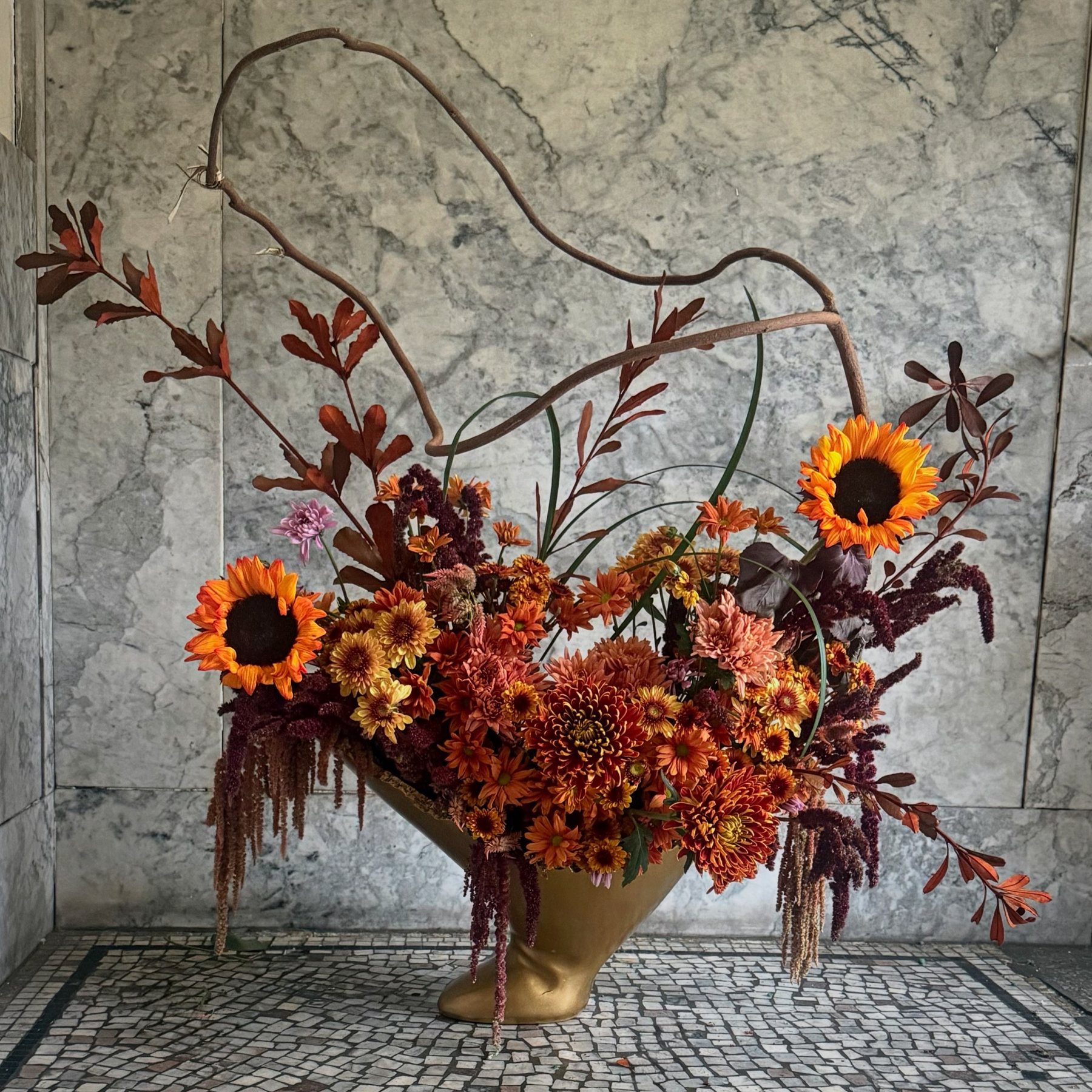 A long contorted vine arcs above chrysanthemums, sunflowers and trailing amaranthus in a tilted gold vessel